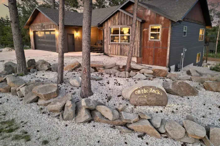 Imagine arriving at "On The Rocks" in Powder House Pass, stepping into the rustic charm of this inviting cabin. Picture the warm lights glowing against the evening sky, the crunch of gravel, and the natural rock garden welcoming you to a peaceful retreat.