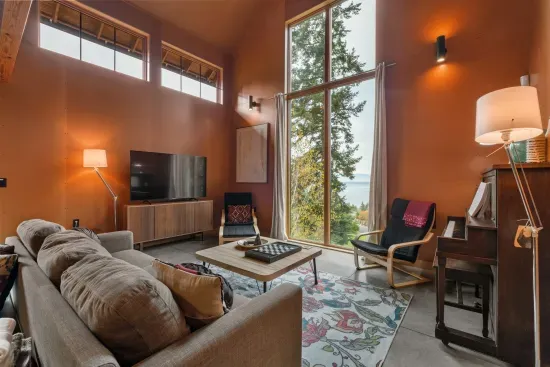 The living room boasts a stunning 14-foot-tall scenic window with views of the water and mountains, along with high ceilings, a smart TV, a piano, and a spacious coffee table perfect for games, drinks, and snacks.
