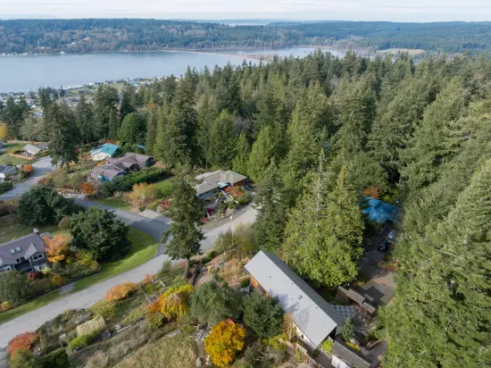 An aerial view showcases the house nestled into the hillside, surrounded by lush woods, with stunning views of Cutlass Cove and the shimmering waters of Puget Sound.