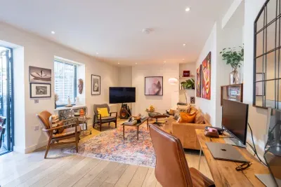 Styled like a private gallery — original art on every wall, a tan leather sofa, classic lounge chair, and mid-century seating in leather and teak. A proper hi-fi speaker system, brass bar cart and shelves of art books make it a room for slow evenings