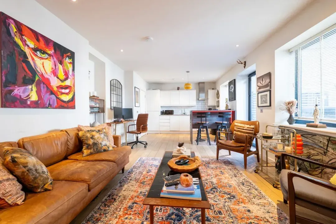 Styled like a private gallery — original art on every wall, a tan leather sofa, classic lounge chair, and mid-century seating in leather and teak. A proper hi-fi speaker system, brass bar cart and shelves of art books make it a room for slow evenings