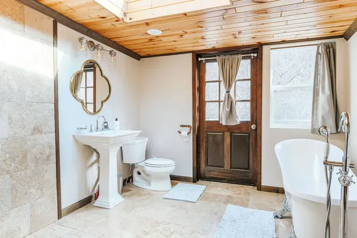 Bright, spacious bathroom featuring a soaking tub, elegant pedestal sink, and natural wood accents.