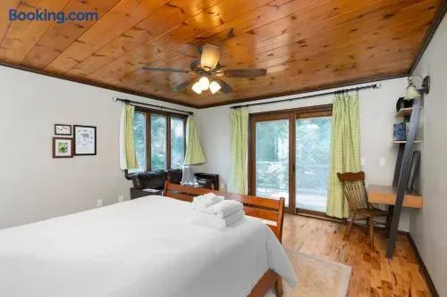 Bright, rustic bedroom with a warm wood-paneled ceiling, large windows and glass doors overlooking the outdoors, a neatly made bed with white linens, green patterned curtains, hardwood floors, and a cozy sitting/work area.