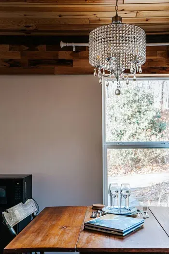 Charming dining area with a sparkling crystal chandelier, rustic wood ceiling, warm wooden dining table, and large window overlooking the surrounding greenery.