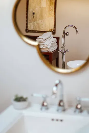Elegant bathroom detail featuring a round gold-framed mirror reflecting the freestanding tub and neatly folded white towels, with the foreground sink softly blurred for a refined, spa-like feel.