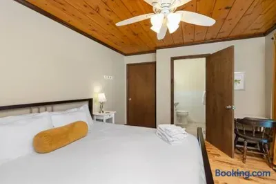 Cozy rustic bedroom featuring a neatly made bed with crisp white linens and a mustard bolster pillow, warm wood-paneled ceiling, ceiling fan, hardwood floors, bedside table with lamp, and an adjoining bathroom.