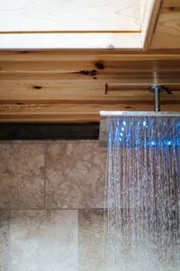 Modern walk-in shower with a sleek rainfall showerhead, illuminated by cool blue lighting against warm wood ceilings and natural stone tile walls.