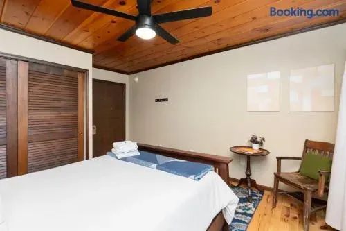 ozy rustic bedroom with a warm wood-paneled ceiling, ceiling fan, large bed with crisp white linens and blue accents, wooden furniture, a comfortable armchair, and a small side table.