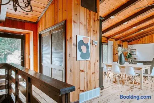 Warm, rustic cabin interior with natural wood walls and exposed beams, connecting a cozy upper landing to a bright dining area with modern white seating and minimalist décor.