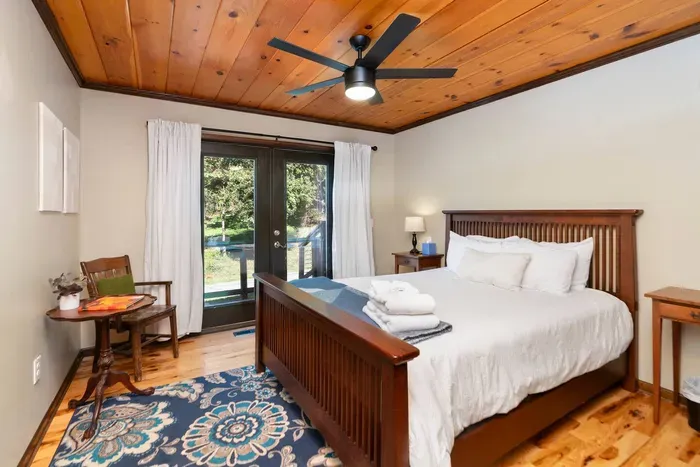 Serene Bedroom: Comfortable king-size bed with fresh linens, warm wood accents, a ceiling fan, and direct access to a private outdoor deck surrounded by nature.