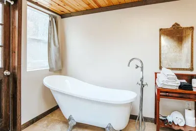Bright, inviting bathroom featuring a freestanding white soaking tub, elegant vintage gold-framed mirror, polished tub faucet, and neatly folded towels on a wooden console.