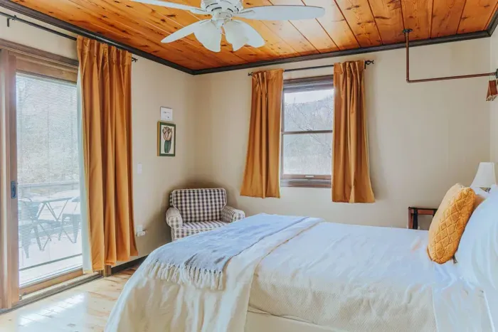 Cozy bedroom with a comfortable bed, natural light, wood-paneled ceiling, and a small sitting area.