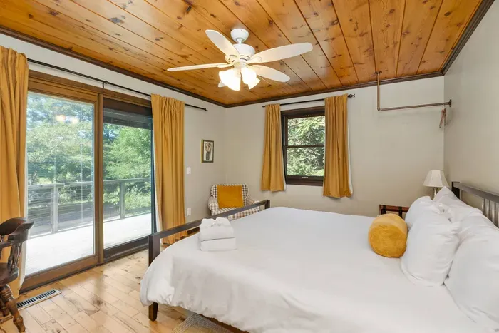Spacious Bedroom: Comfortable bed with fresh linens, warm wood accents, ceiling fan, and large windows offering peaceful views of the surrounding greenery.