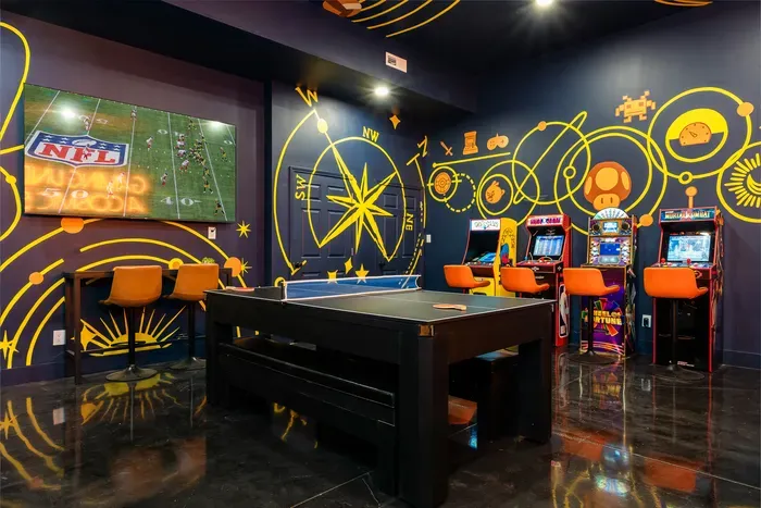 Game night anchor. Ping pong front and center, the arcade lineup waiting along the wall, the TV keeping the game on in the background. The room everyone ends up in.