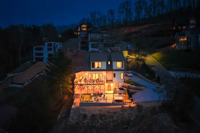 The house lit like a lantern over the Blue Ridge dark, decks stacked above the patio below. Fifteen minutes from downtown Asheville, the rest of the world an easy drive away.