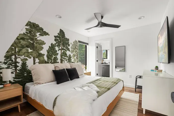 Upstairs king bedroom with striking forest mural, ceiling fan, TV, and ensuite bathroom. A cozy retreat with modern design details and plenty of natural light.