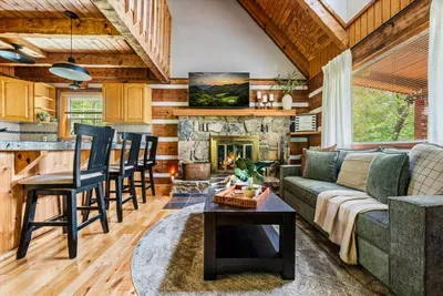 Open-concept great room with wood-burning fireplace, dining for six, cathedral log ceiling, and mountain light pouring through every window. This is the heart of it.
