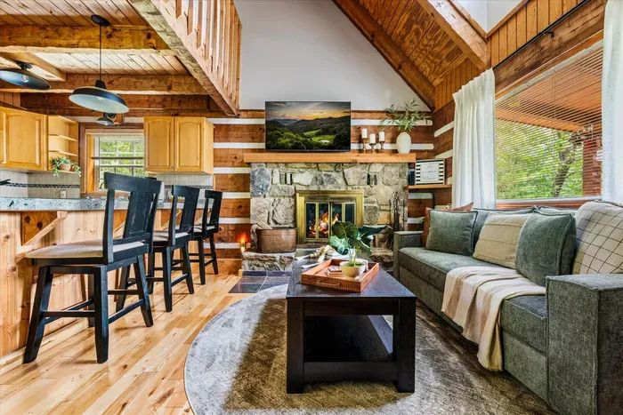 Open-concept great room with wood-burning fireplace, dining for six, cathedral log ceiling, and mountain light pouring through every window. This is the heart of it.