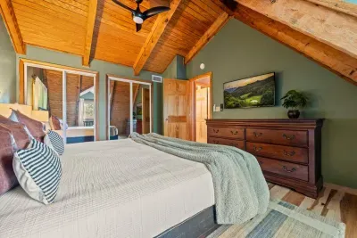 A peaceful A-frame bedroom in forest green — vaulted beams, a solid dresser, and a slow ceiling fan overhead. Pull the curtains, settle in, and let the woods keep things quiet.
