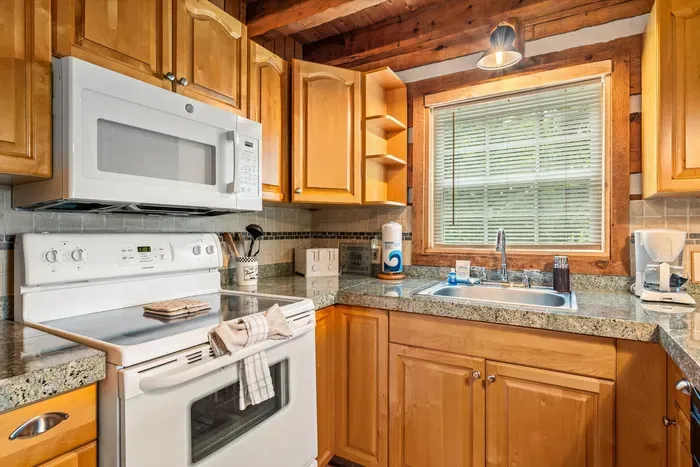 Full-size range, microwave, and granite prep space framed by log walls and a window over the sink — the kind of kitchen that turns a rainy day into a from-scratch dinner. Bring the recipes.