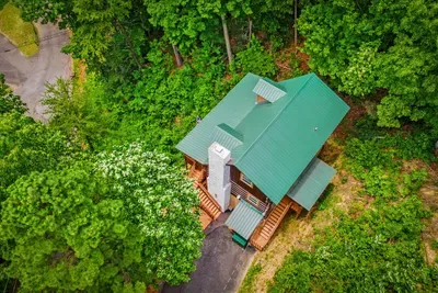 Your mountain escape from above — green metal roof, private wraparound deck, and a full canopy of Blue Ridge hardwoods holding you in from the world.