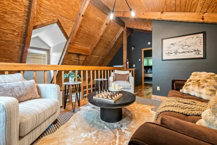 Up in the A-frame loft, a relaxed lounge gathers around the rug — soft seating, a glowing mountain wall, and the whole cabin humming below. The retreat-within-a-retreat upstairs.