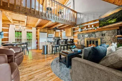 A soaring open-concept great room anchored by a floor-to-ceiling stone fireplace — vaulted wood ceilings, a loft above, and the full kitchen just steps away. The heart of the whole cabin.