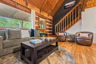 Open log-walled living room with a staircase climbing to the loft, deep seating, and windows full of green — the gathering space where the whole group lands. Room to sprawl, light to spare.