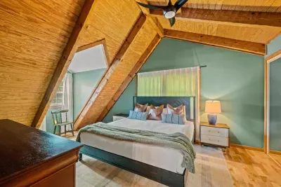 Sleep beneath the A-frame peak — sage-green walls, layered bedding, and a window framing the treetops. The slanted-ceiling room guests always ask for first.