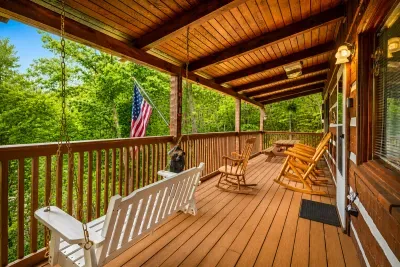 Rocking chairs lined up on a covered porch, flag snapping in the breeze, woods rolling out past the rail — claim your chair and let the afternoon stretch. The spot everyone fights over.