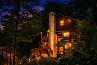 Cedar and stone rise through the Blue Ridge hardwoods — two stories of handcrafted mountain cabin, soaring chimney, upper deck, and summer green all around.