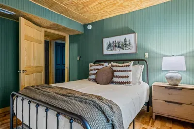A crisp white bed framed in black iron, pine-forest art overhead, and a low-glowing nightstand lamp — this guest room invites the deepest sleep of the whole trip.