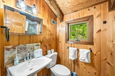 A wood-paneled bathroom with a pedestal sink and a window framing the green outside — fresh towels hung and forest light pouring in. Cabin simplicity, done right.