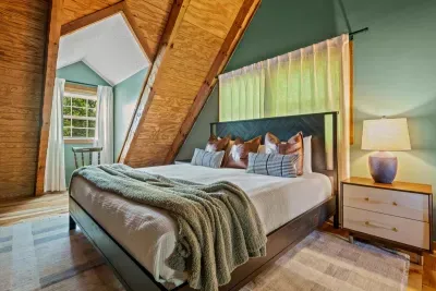 An A-frame bedroom bathed in green light — a soft throw across the bed, a lamp glowing on the nightstand, woods just beyond the glass. Where mountain mornings start slow.
