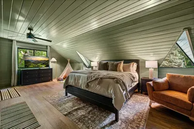 Loft-level bedroom with green shiplap, an A-frame slope overhead, and a reading chair by the window. Tucked-away privacy with the treetops right at eye level.