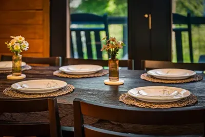 Fresh flowers, woven chargers, and a table set and waiting — the thoughtful touches that greet you the moment you sit down. Cabin dining with the deck and trees in view.