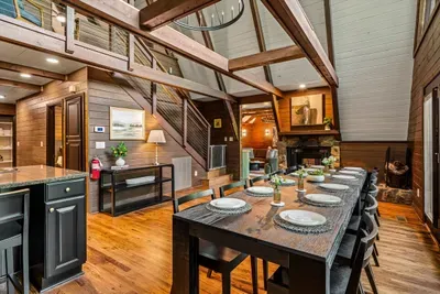 Long farmhouse table set beneath the loft staircase, fireplace glowing at the far end. Seats the whole group for big breakfasts and lingering dinners with the forest just outside.