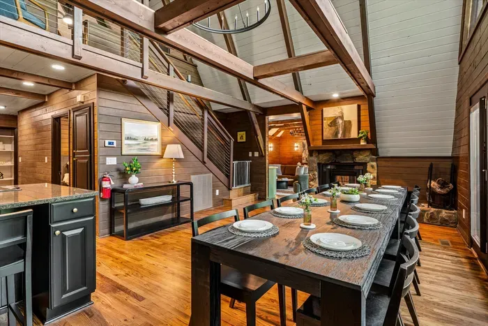 Long farmhouse table set beneath the loft staircase, fireplace glowing at the far end. Seats the whole group for big breakfasts and lingering dinners with the forest just outside.