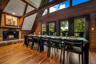 Long harvest table for twelve beside a crackling stone fireplace, framed by soaring A-frame windows. Family dinners hit different when the forest is your backdrop.