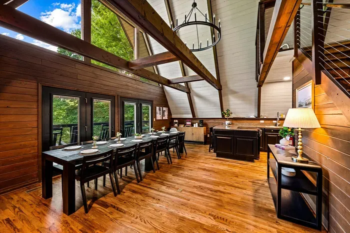 Harvest table for the whole party beneath towering A-frame windows and a wrought-iron chandelier. Long dinners, big laughs, forest glowing green through every pane.