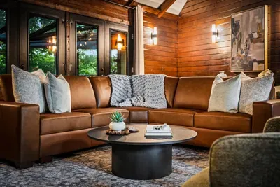 Open great room wrapped in leather seating and windows to the woods, glowing under a wall of Edison light. The gather-everyone hub of the whole cabin.
