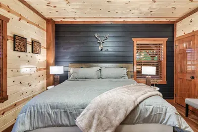 King bedroom with deep navy walls, gray linen bedding, and stag art overhead — moody mountain retreat energy that makes 9pm feel like the perfect bedtime.