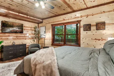 Bright knotty pine bedroom with dual windows, a dresser full of drawer space, and mountain landscape art — unpacked and at home within minutes of arriving.