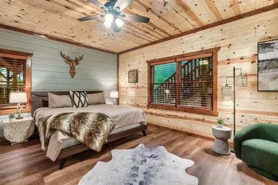 King bedroom with hardwood floors, cowhide rug, antler mount, and a ceiling fan for cool mountain nights — authentic cabin character from floor to rafter.