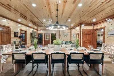 Grand dining table set for 12 under a statement chandelier and vaulted knotty pine — the cabin's heartbeat. Every group needs a table big enough for the whole story.