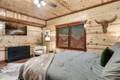 Fireplace bedroom with a longhorn above the mantle, vaulted pine ceiling, and gray linens — rugged mountain character meets the warmth of a crackling private fire.