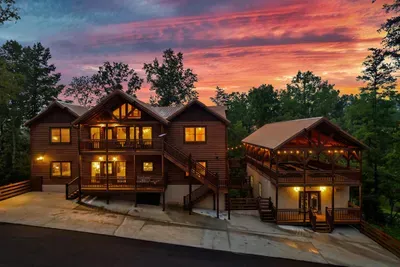 Two cabin buildings strung with warm lights under a sweeping rose-gold Smoky Mountain sky — the kind of property photo that makes you stop scrolling and start planning.