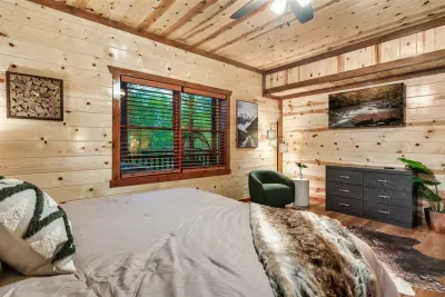 Warm knotty pine bedroom with a fur-throw king bed, green accent chair, and tree-framed windows — the kind of room that slows you down in the best possible way.