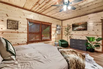 Airy king bedroom under vaulted knotty pine with a ceiling fan, lush plant accent, and warm cabin light — bright, breathable, and ready to feel like yours from day one.