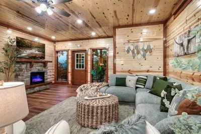 Open-plan great room with a stone fireplace, oversized gray sectional, and wicker ottoman — gather here after a long day in the mountains and let the evening take care of itself.